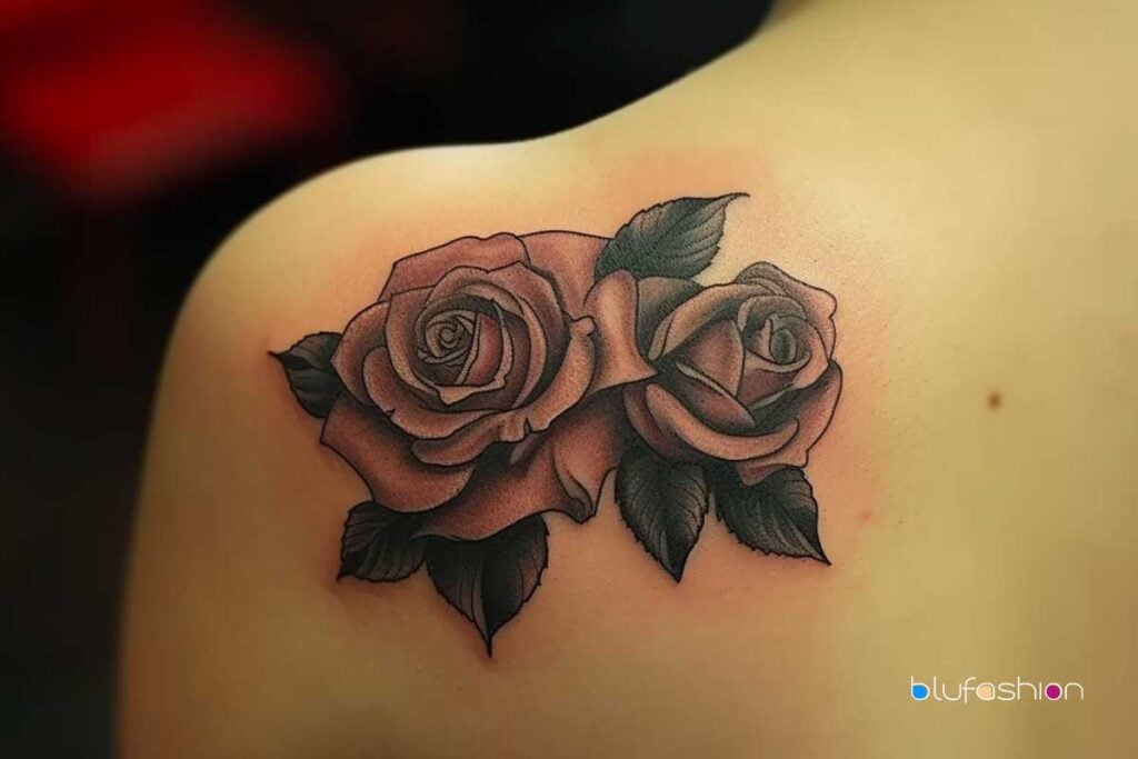 Par de tatuajes de rosas con sombreado rosa suave y hojas oscuras en un hombro liso.