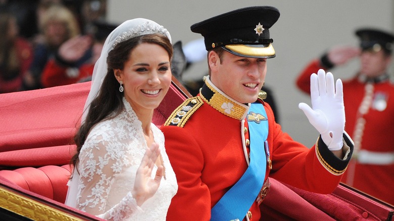 Kate Middleton sentada en un carruaje junto al príncipe William. Lleva un vestido de novia blanco de encaje con cuello alto, tiara y velo. William lleva un uniforme rojo de capitán y una gorra de forraje.