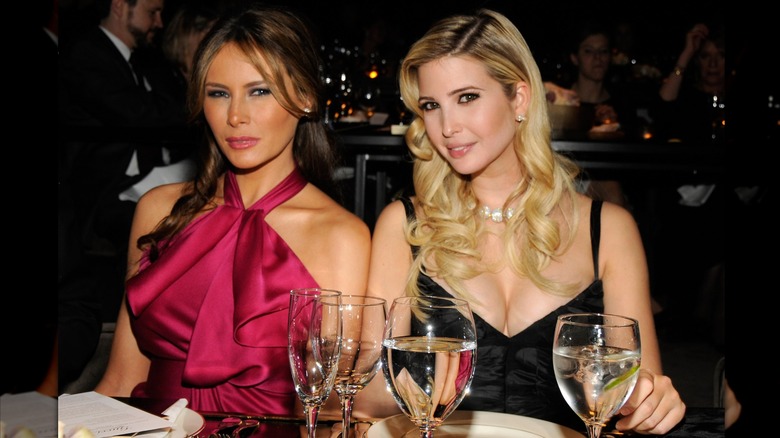 Melania e Ivanka Trump se sientan una al lado de la otra en una mesa. Melania lleva un vestido sin mangas de color baya con pliegues. Ivanka lleva un vestido negro de tirantes finos.