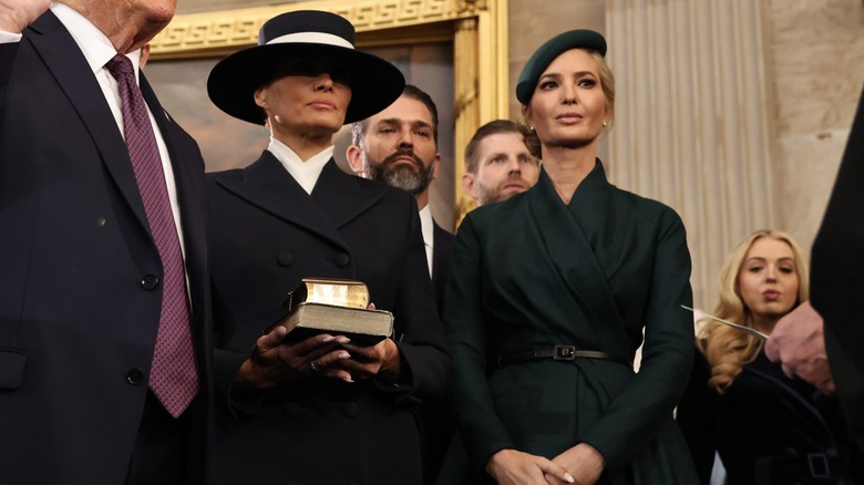Melania lleva un abrigo azul marino con hombreras, una blusa blanca de cuello alto y un sombrero azul marino de ala ancha con una cinta blanca. Sus ojos están cubiertos por el sombrero. Ivanka lleva un abrigo cruzado de color verde oscuro con una falda a juego y una boina verde inclinada hacia un lado.