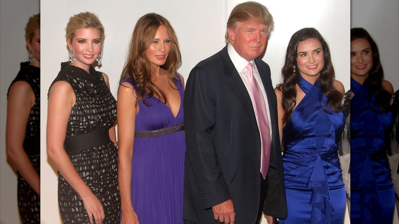 Ivanka y Melania Trump posan junto a Donald Trump y Demi Moore, una mujer de piel clara y cabello castaño ondulado. Ivanka lleva un vestido de lunares grises y negros con un cinturón grueso. Melania lleva un vestido morado de cintura imperio.
