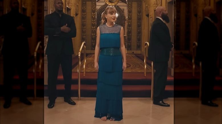 Swift en el pasillo de un gran hotel. Lleva un vestido azul con varias capas de flecos. Su cabello está recogido en una cola de caballo.