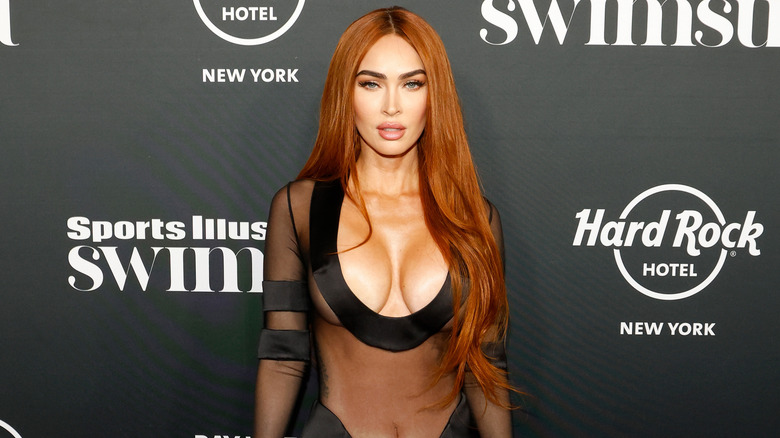 Megan Fox lleva un vestido negro transparente y escotado y tiene el pelo rojo con raya en medio.