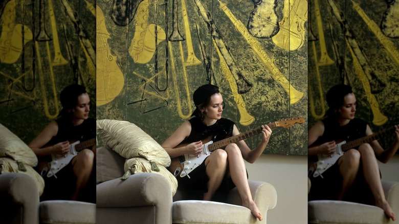 Winona Ryder tocando la guitarra en una silla frente a una colorida obra de arte.