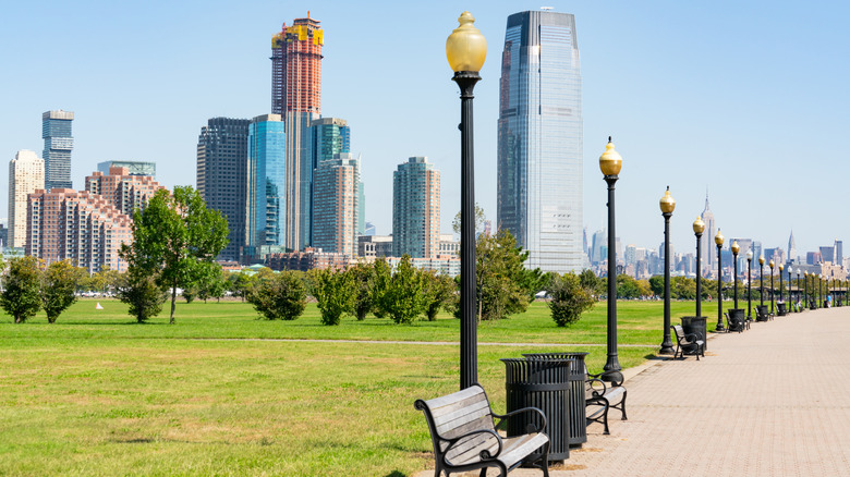 Amplia zona verde bordeada de farolas y bancos de madera, los horizontes de Jersey City y Manhattan en la distancia, Liberty State Park