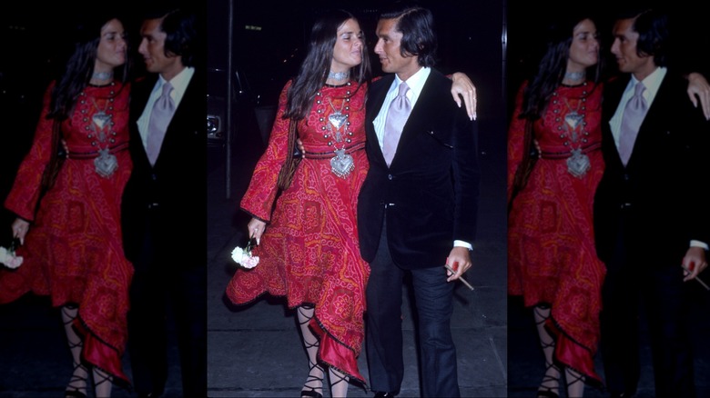 Ali MacGraw con un vestido rojo, sandalias de tiras y un collar grande con el brazo alrededor de Robert Evans con traje.