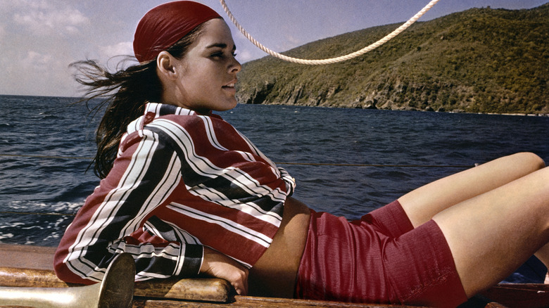 Ali MacGraw recostado en un velero con un pañuelo rojo, camisa a rayas y pantalones cortos rojos.