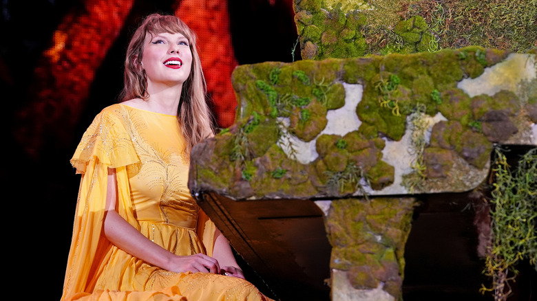 Taylor Swift sonríe a la multitud mientras lleva un vestido amarillo y está sentada frente a un piano cubierto de musgo.