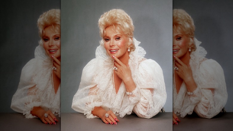 Eva Gabor posa con la mano en la cara luciendo una blusa de encaje abullonada y un largo collar de perlas.