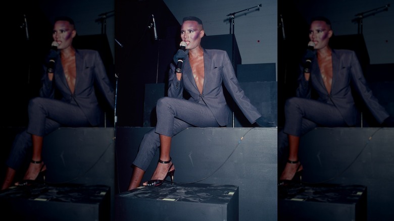 Grace Jones vistiendo un traje gris con escote en V profundo mientras se sentaba y cantaba en el escenario.