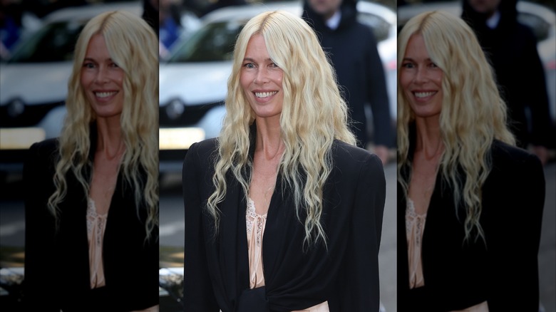 Claudia Schiffer sonriendo con un top blanco y un abrigo negro con cabello rubio con ondas de sirena.