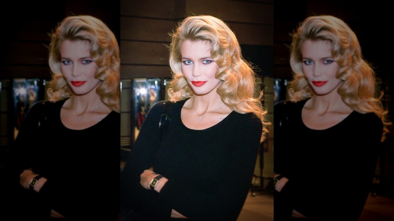Claudia Schiffer posa con un top negro con escote redondo, lápiz labial rojo, brazos cruzados y cabello rubio rizado y desgastado.