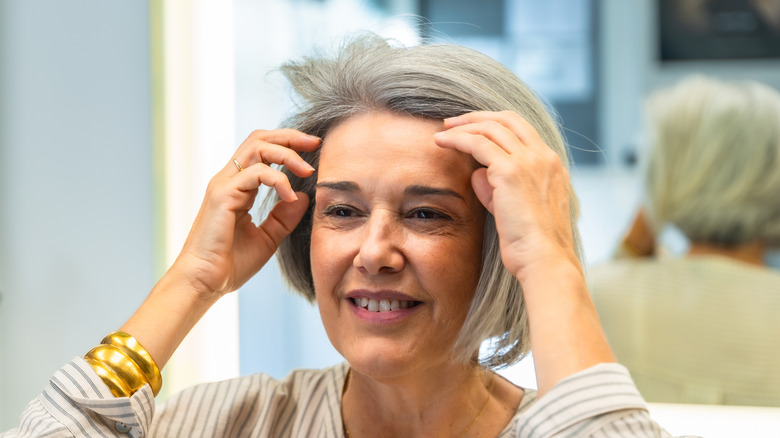 Mujer madura sonriendo y arreglando su cabello con un espejo detrás de ella.