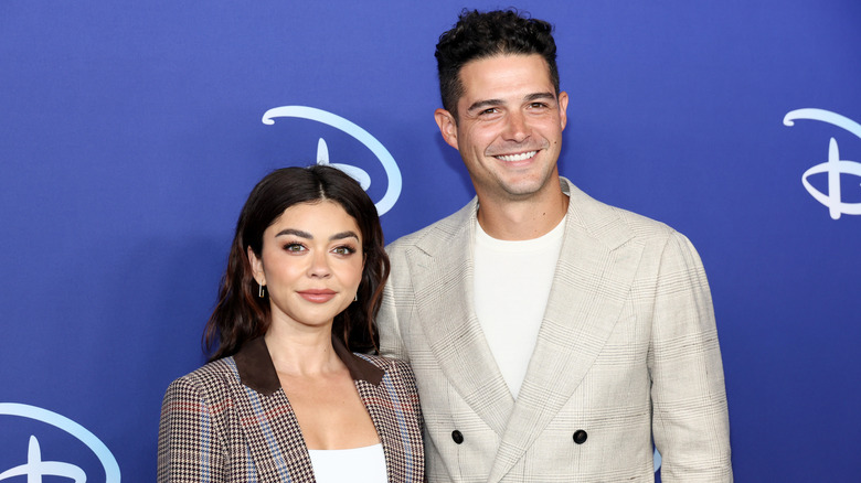 Sarah Hyland con una chaqueta a cuadros marrón posando junto a Wells Adams con una chaqueta color crema
