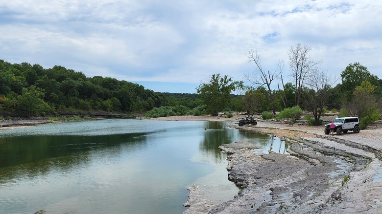 Un vehículo todoterreno y un jeep sentados en la orilla rocosa junto a un río ancho y poco profundo con árboles verdes en la orilla opuesta