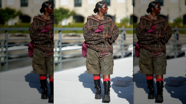 Una mujer que vestía una chaqueta de tartán marrón y verde oliva, pantalones cortos color oliva y botas negras.