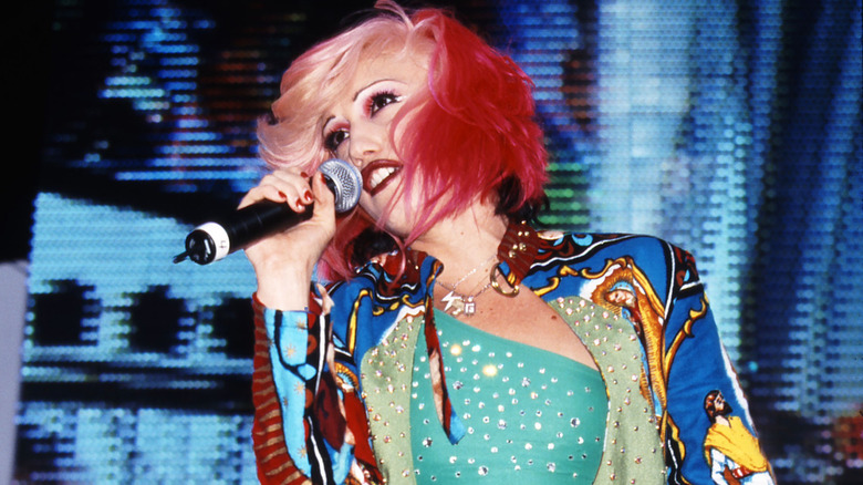 Gwen Stefani de No Doubt actuando con cabello rosado y rubio vistiendo una camisa turquesa y una chaqueta colorida.