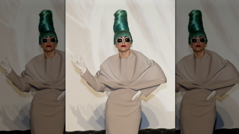 Lady Gaga actuando con un vestido beige con un peinado de colmena verde azulado y gafas de sol.