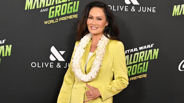 Tia Carrere lanzando un cartel de shaka en un evento de alfombra roja vistiendo un traje amarillo claro y un lei.
