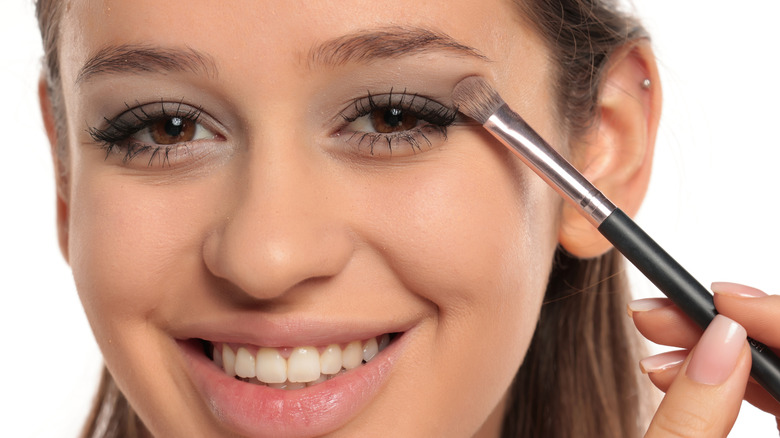 Mujer sonriendo y mirando a la cámara mientras aplica sombra de ojos con un pincel pequeño