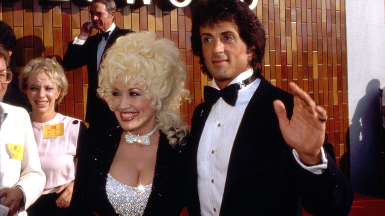 Dolly Parton en 1984 posando con Sylvester Stallone vistiendo un top plateado brillante con pajarita a juego y una chaqueta negra brillante.