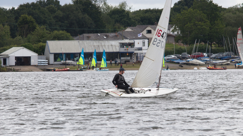 Un hombre viajando en un velero en Pennington Flash con el club náutico y los autos detrás de él.