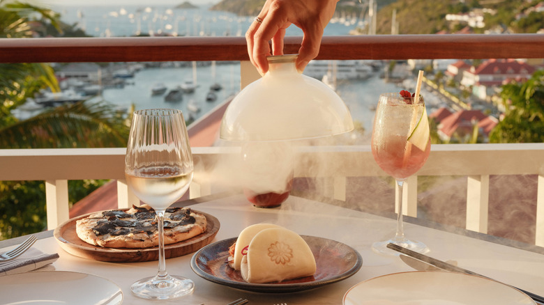 Una mano levantando una cúpula de cristal llena de humo de un plato que revela bollos bao en una mesa de comedor con vistas al mar Caribe en el restaurante Beefbar en Fouquet's Saint-Barth.