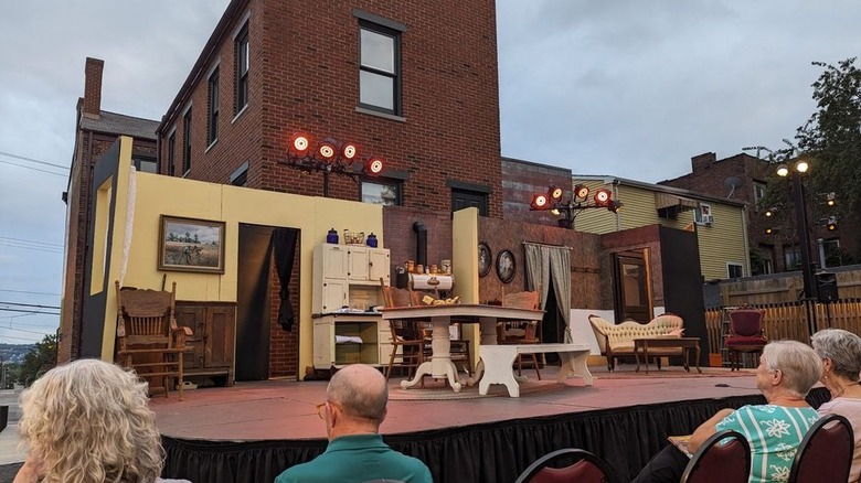 Escenario en el patio trasero de la Casa August Wilson, con una cocina con paredes amarillas y una sala de estar con paredes marrones frente a un edificio alto de ladrillo y miembros de la audiencia sentados frente a él, bajo un cielo nublado.
