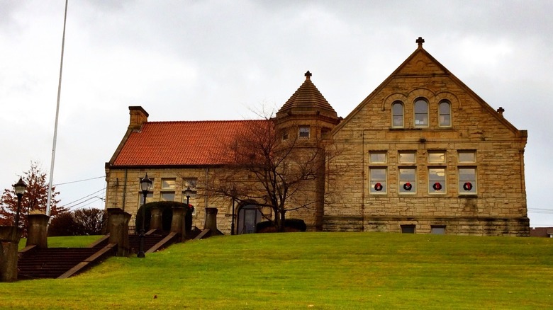 Biblioteca Carnegie de McKeesport, un edificio de piedra marrón con techo rojo y torre octogonal, con coronas en cuatro de las ventanas y una escalera de piedra que conduce a través de hierba verde, bajo un cielo nublado