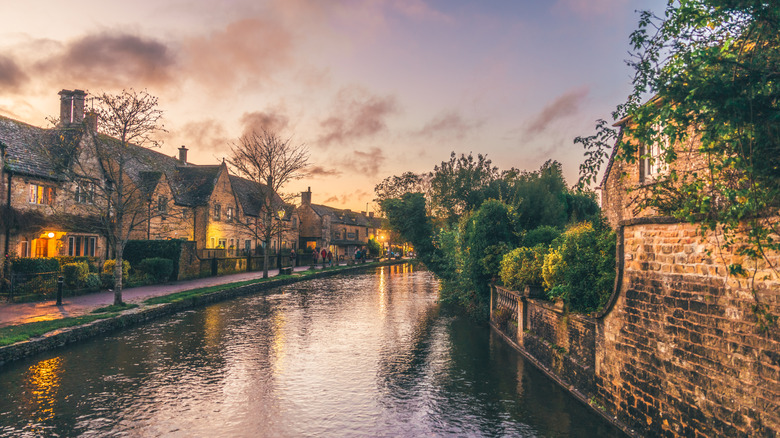 Un canal que atraviesa Bourton-on-the-Water, con casas iluminadas flanqueándolo a la izquierda, bajo un cielo colorido al atardecer