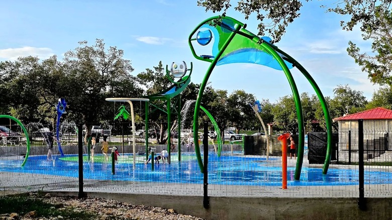 Varios niños juegan en una zona de chapoteo en Brushy Creek Lake Park en Texas, que cuenta con chorros de agua azules y verdes y fuentes rociadoras.