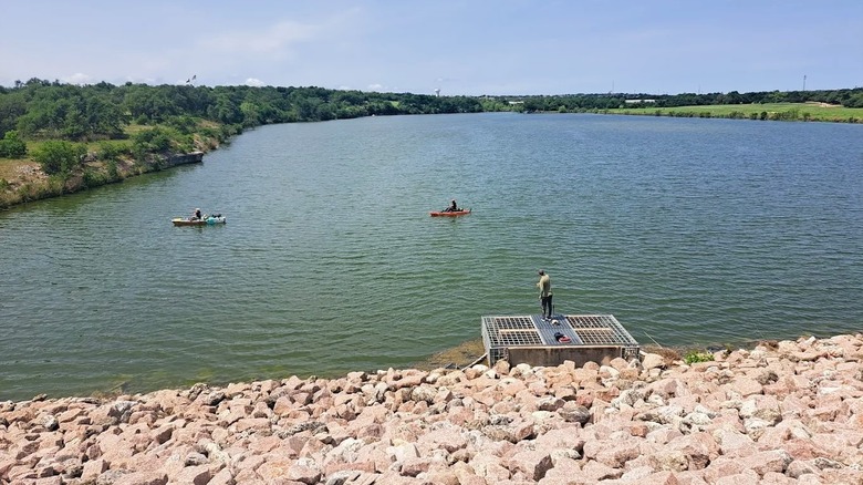 Visitantes pescando en un lago tranquilo en Brushy Creek Lake Park cerca de Austin, Texas, que cuenta con un muelle, un terraplén rocoso y vegetación.