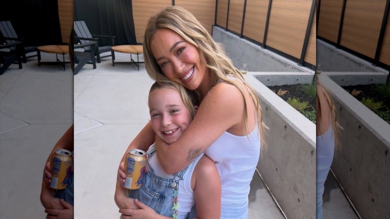 Hilary Duff lleva su cabello rubio suelto en ondas de sirena mientras usa una camiseta sin mangas blanca y posa con su hija Banks durante su 