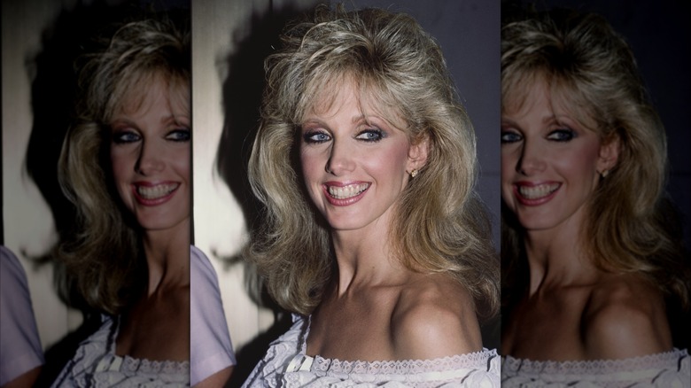 Morgan Fairchild en 1984 sonriendo con un vestido con hombros descubiertos y flequillo esponjoso.