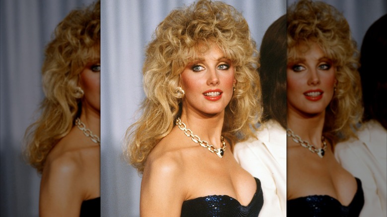 Morgan Fairchild en 1982 en un evento formal luciendo un vestido brillante y un collar de cadena con un voluminoso cabello rizado.