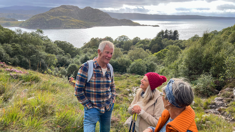 Tres excursionistas sonríen y charlan en un claro cubierto de hierba sobre un lago en un día nublado en Escocia