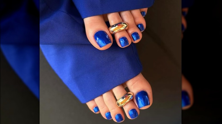 Una pedicura con esmalte de uñas azul cromado y anillos dorados en los pies.