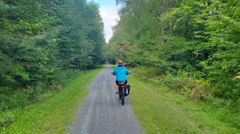 Una persona con una chaqueta azul y un sombrero negro y naranja de ala ancha anda en bicicleta por el Ghost Town Trail, con árboles y pasto creciendo a ambos lados