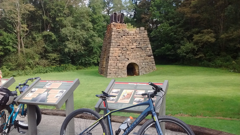 Eliza Furnace, una estructura de piedra trapezoidal con tubos de metal en la parte superior, rodeada de césped con árboles al fondo y carteles interpretativos en primer plano con bicicletas azules apoyadas en ellos.