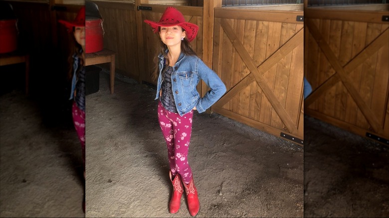 Skywalker Hughes sonriendo con un sombrero de vaquera rojo y botas con leggings con estampado floral rosa y una chaqueta vaquera.