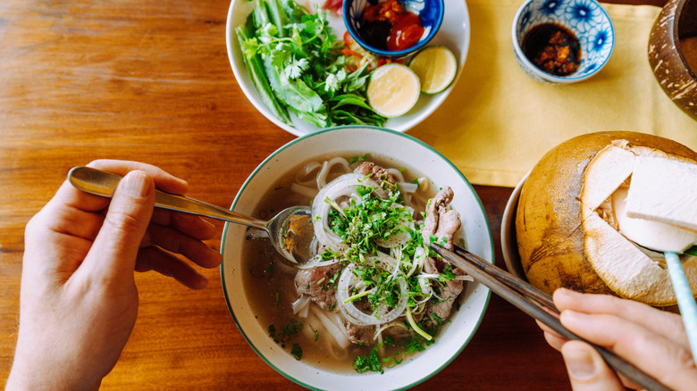 Vista superior de un par de manos sobre un plato de pho con tazones de condimentos y una bebida de coco
