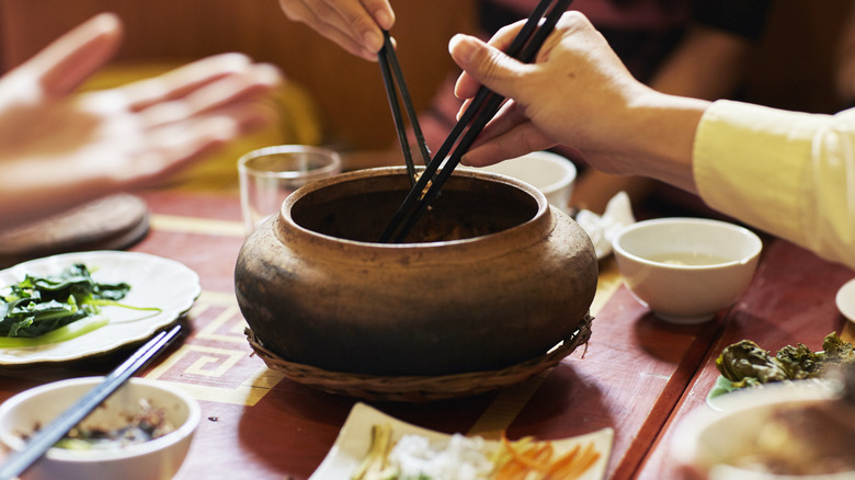 Dos pares de palillos sumergidos en una olla de barro comunitaria con pequeños platos y tazones al fondo