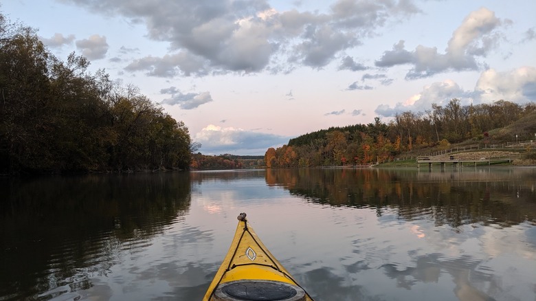 Lago Cross Creek en otoño, con una punta de kayak amarilla en primer plano sobre el agua rodeada de árboles en otoño con un muelle de madera a lo largo del lado derecho del marco, bajo un cielo nublado al atardecer
