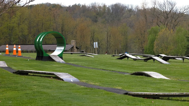 Parque de bicicletas para niños en Cross Creek Park, con rampas de madera, un tubo verde y conos de tráfico de color naranja rodeados de césped, con un pabellón y árboles de invierno al fondo