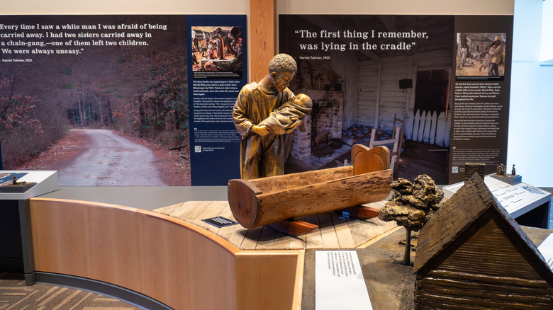 Exposiciones dentro del Centro de Visitantes, con una estatua de bronce de un hombre sosteniendo a un bebé en el centro detrás de una cuna de madera, con una pequeña casa modelo en primer plano y carteles con palabras detrás