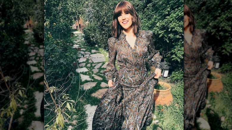 Se ve a Erin Krakow sosteniendo la falda de un vestido estampado fluido frente a un pintoresco fondo verde.