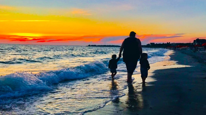 Siluetas de un padre caminando de la mano de dos niños a lo largo de Old Silver Beach al atardecer, con una puesta de sol de color naranja resplandeciente
