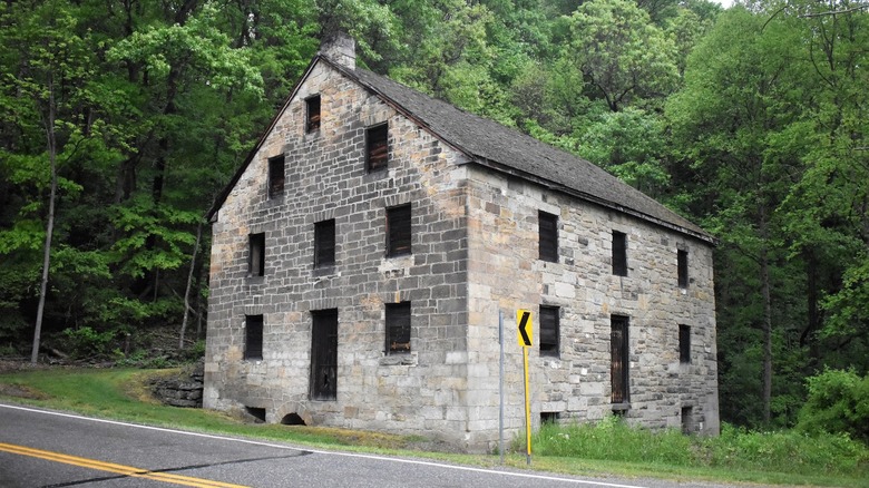 Hambleton's Mill, un edificio de piedra con ventanas y puertas tapiadas junto a una carretera, con un cartel amarillo al lado y árboles al fondo