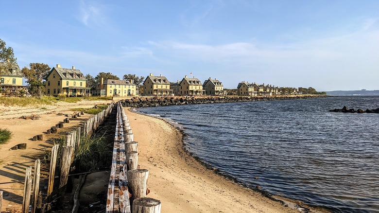 Orilla arenosa con una valla de madera y un malecón de roca a la izquierda y agua azul a la derecha
