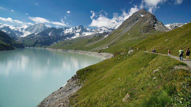 Los turistas caminan por un sendero a lo largo de las orillas del Lac des Dix en los Alpes Peninos durante un día soleado con las montañas al fondo.